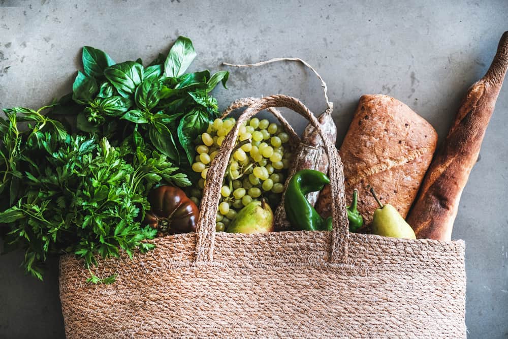 Flat-lay of healthy grocery shopping eco-friendly bag with vegetables, fruit, greens, herbs, bread and sausage over concrete background, top view. Local farmers market, shopping mall concept